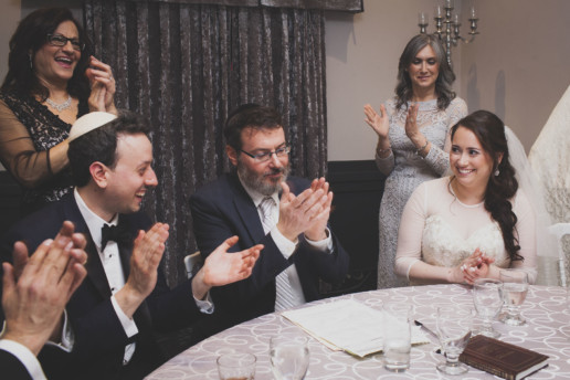 Mother of the bride and mother of the groom and the couple and rabbi are all clapping and smiling when signing the ketubah for a Jewish wedding