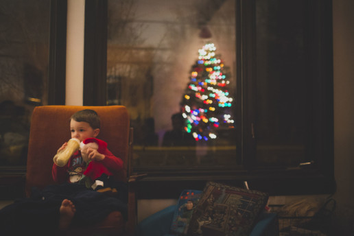 A little boy drinks from his bottle at night while holding his Charlie Brown Peanuts doll and sits in a big orange arm chair with a reflection of a Christmas tree in the window