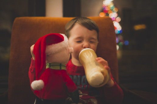 A little boy drinks his bottle at night with his eyes squeezed tightly closed while sitting in an orange chair and holding his Charlie Brown Peanuts doll with the reflection of the Christmas tree in the window behind him in Pointe-Claire Quebec
