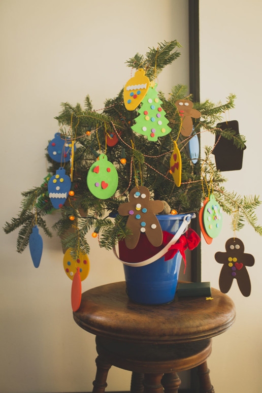 A child's Christmas tree is decorated with foam ornaments and sits on a wooden stool in the warm window light