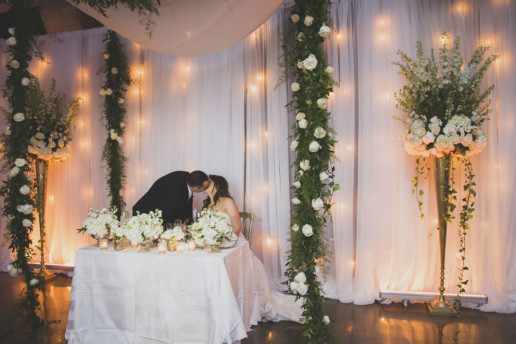 Couple kiss under their wedding canopy at Gare Viger in Montreal amid white flowers and greenery for a neutral colour palette