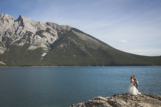 bride and groom stand on a rock in front of Lake Minnewanka in Banff at destination wedding