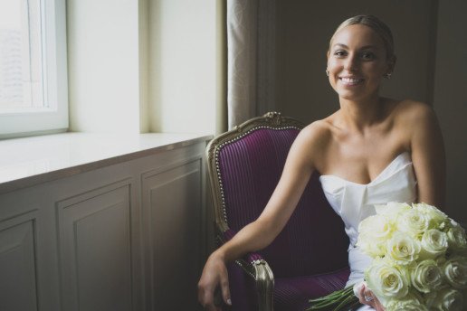 A bride smiles while holding her bouquet of white roses while sitting in a purple armchair in her suite at teh Ritz-Carlton Montreal
