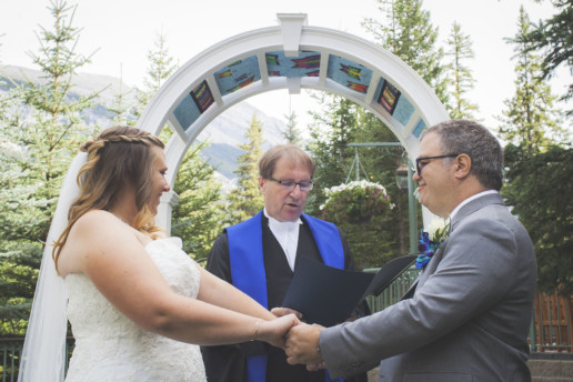 man and woman hold hands during wedding ceremony in front of the officiant under wedding arch outside in Banff