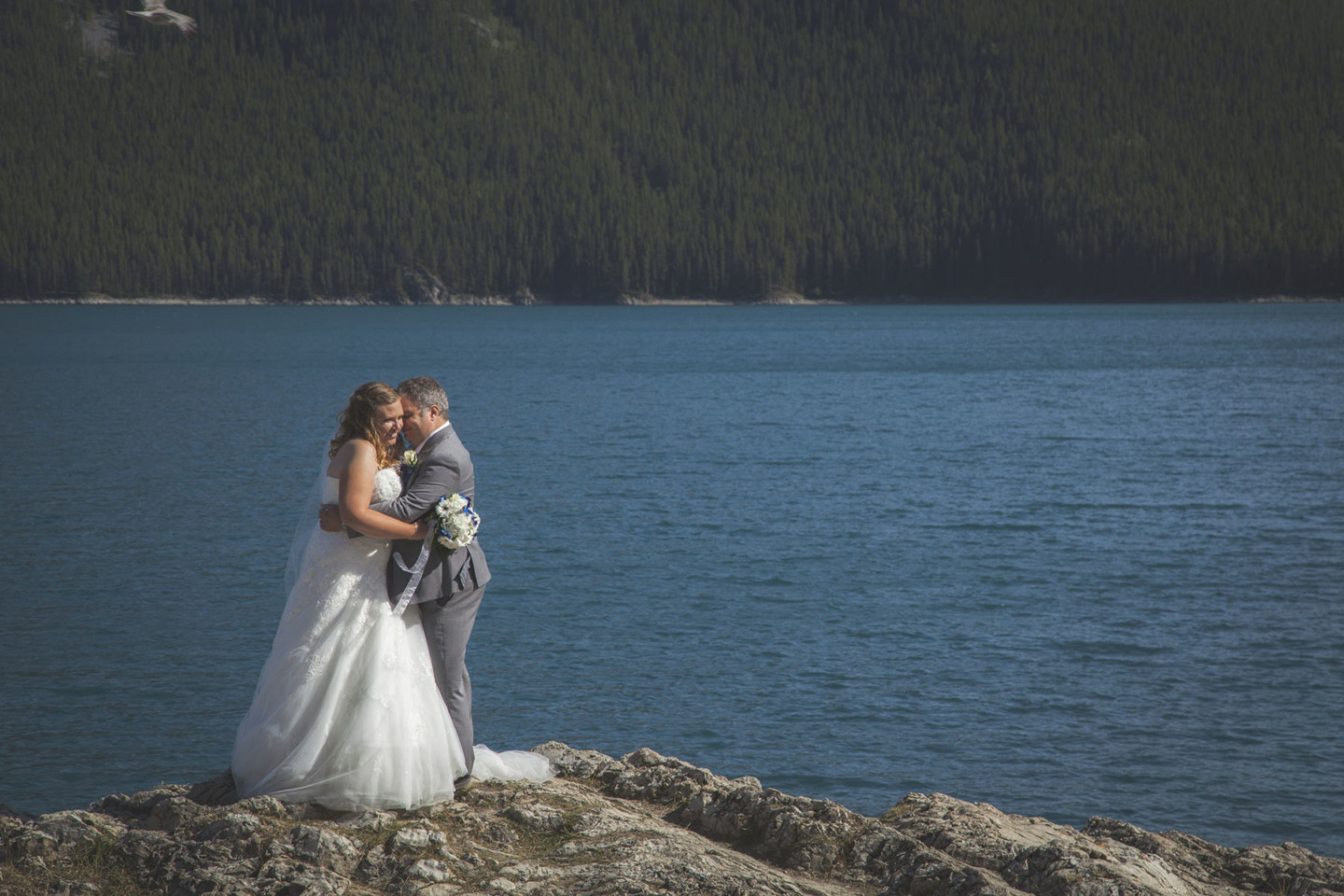 Bride and groom embrace while standing on a rock at Lake Minnewanka in Banff Alberta