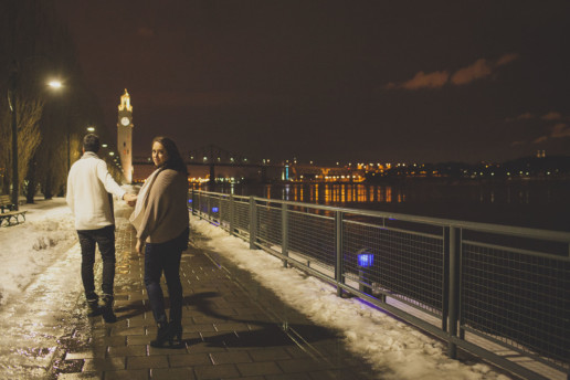 during their engagement shoot a woman looks back while walking hand in hand with a man along the clock tower pier in the old port of Montreal