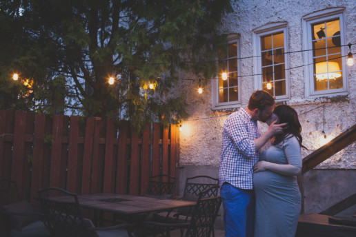 man and woman share a kiss in their backyard at dusk under market lights while she rests her hand on her pregnant belly