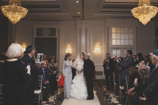 Father of the bride kisses her when she walks down the aisle as guests look on and take pictures on iPhones and iPads