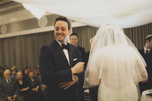 Groom smiles during wedding ceremony as bride circles him under the chuppah