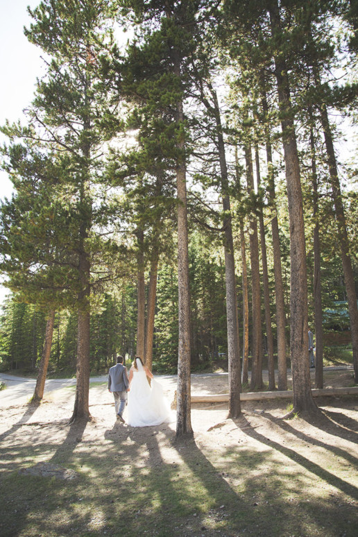 bride and groom walk hand in hand into the distance into the forest as the sun shines through the trees in Banff at destination wedding