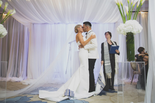 A couple shares their first kiss during their Jewish wedding ceremony while the rabbi smiles and looks on under the chuppah in the Gold room