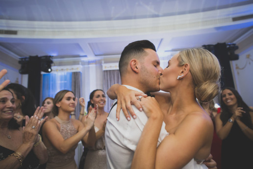 a couple share a kiss on the dance floor with guests clapping and dancing around them in the Oval Room in Montreal
