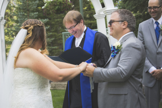 The groom and officiant laugh during a wedding ceremony while he holds the bride's hand in Banff