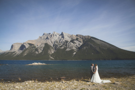 Bride and groom hold hands in front of a mountain at Lake Minnewanka in Banff Alberta