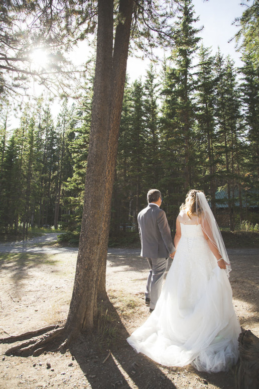 Bride and groom walk hand in hand away from the camera into the setting sun in a forest in Alberta